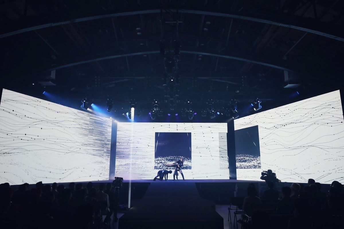 The folded wall lit white with fine line work across all three planes, camera operators silhouetted in front of it.