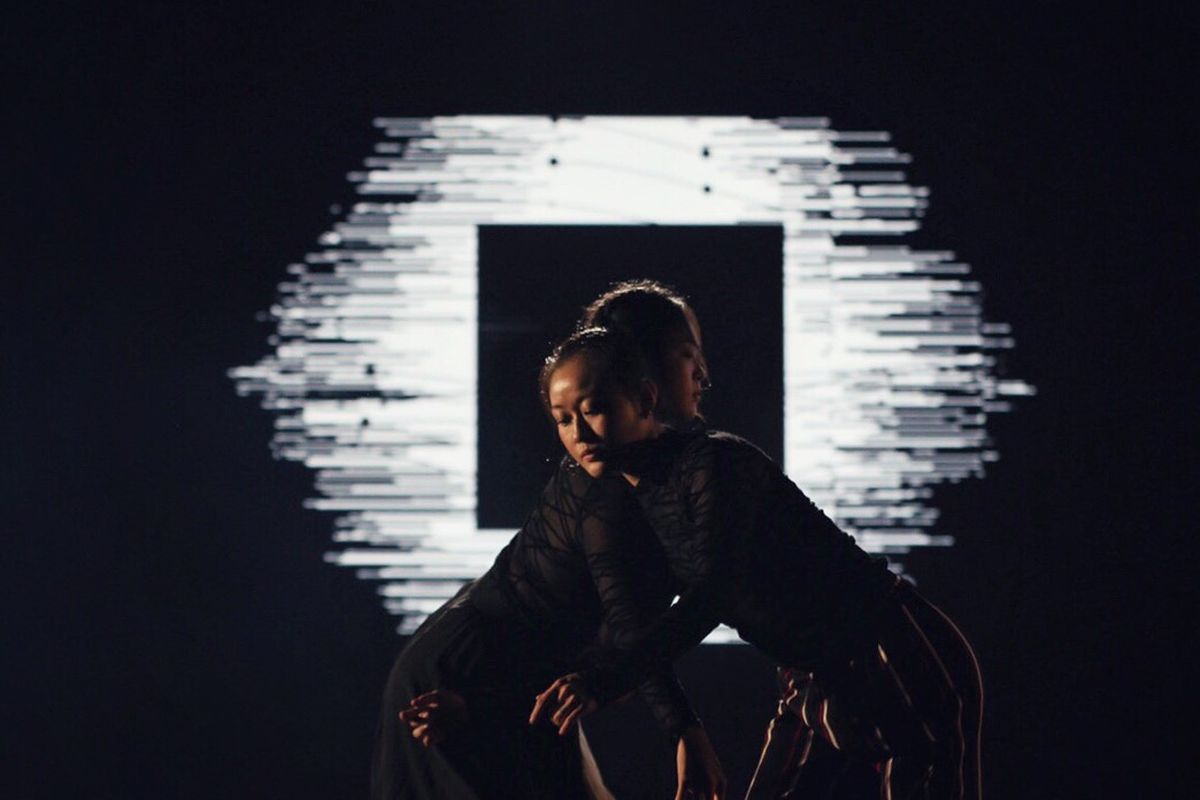 A dancer in black kneeling in front of a black square on the wall, a ragged burst of white particles radiating from behind it.