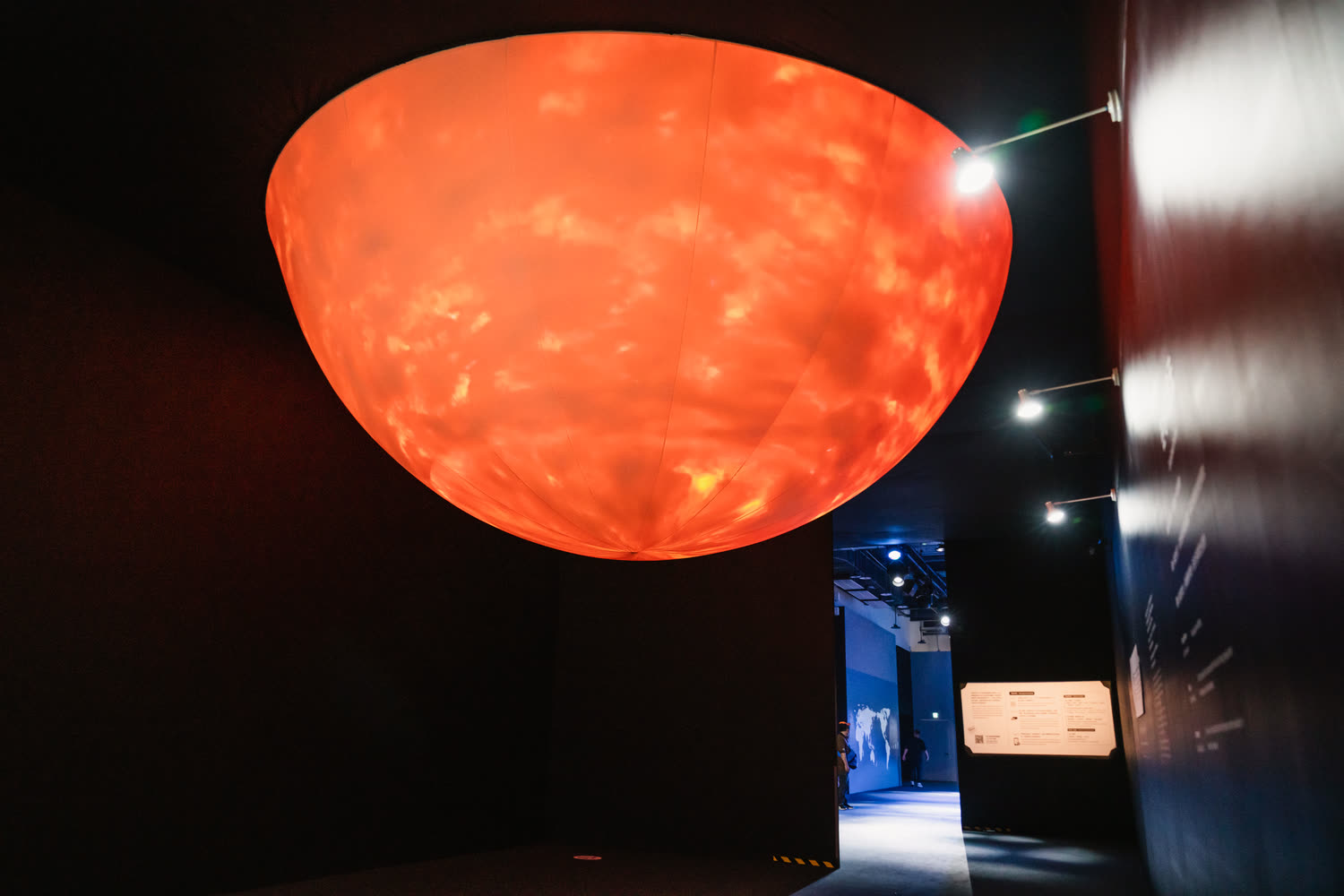 The sphere lit orange from within, seen across the dark room with the lit gallery beyond it.