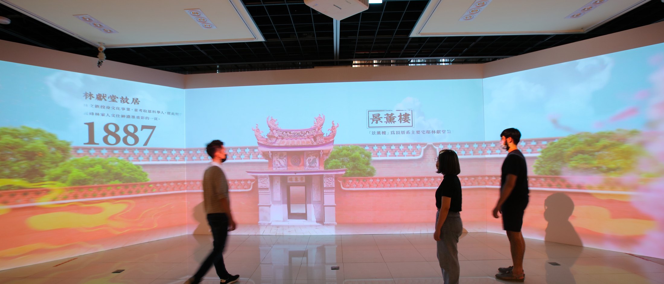 Three visitors standing in a dark room whose walls carry a projected red-brick courtyard wall and ornate gate, dated 1887, the image continuing across the corners and reflecting in the polished floor.